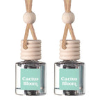 Two matching reed diffuser bottles with clear glass bodies, mint green labels reading 'Cactus Bloom,' beige wooden stoppers, and tan hanging cords on a white background.