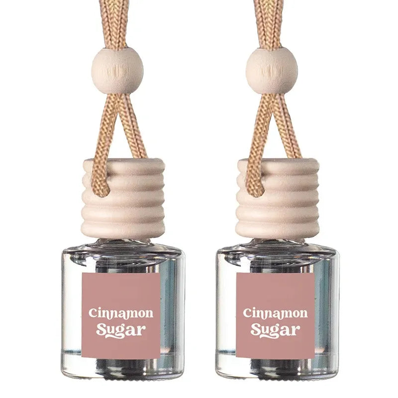 Two clear glass reed diffuser bottles with pink labels reading 'Cinnamon Sugar,' beige wooden stoppers, and tan hanging cords on a white background.