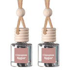 Two clear glass reed diffuser bottles with pink labels reading 'Cinnamon Sugar,' beige wooden stoppers, and tan hanging cords on a white background.