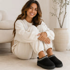 Woman relaxing indoors wearing black Cozy Fleece-Lined House Slippers with a soft suede upper, plush fleece lining, and cushioned platform sole.