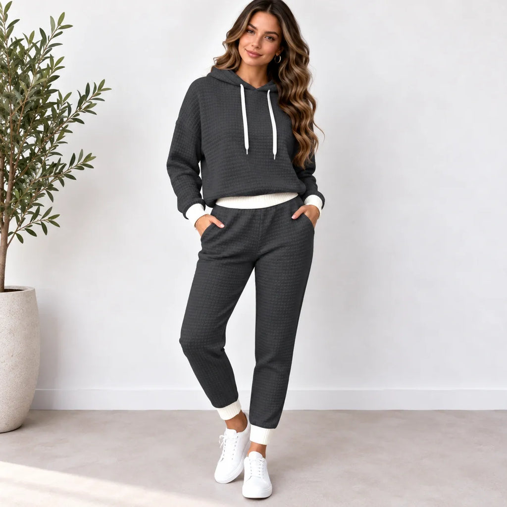 A woman modeling a dark gray waffle-knit two-piece loungewear set consisting of a pullover hoodie and matching jogger pants, styled with white drawstrings, white undershirt hem, white pants cuffs, and white sneakers in a bright indoor setting.