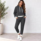 A woman modeling a dark gray waffle-knit two-piece loungewear set consisting of a pullover hoodie and matching jogger pants, styled with white drawstrings, white undershirt hem, white pants cuffs, and white sneakers in a bright indoor setting.