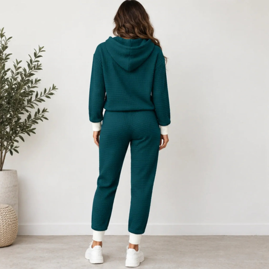 A woman viewed from behind wearing a matching dark teal waffle-knit lounge set consisting of a hooded sweatshirt and jogger pants, styled with white socks and sneakers in a bright room with a potted plant.