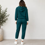 A woman viewed from behind wearing a matching dark teal waffle-knit lounge set consisting of a hooded sweatshirt and jogger pants, styled with white socks and sneakers in a bright room with a potted plant.