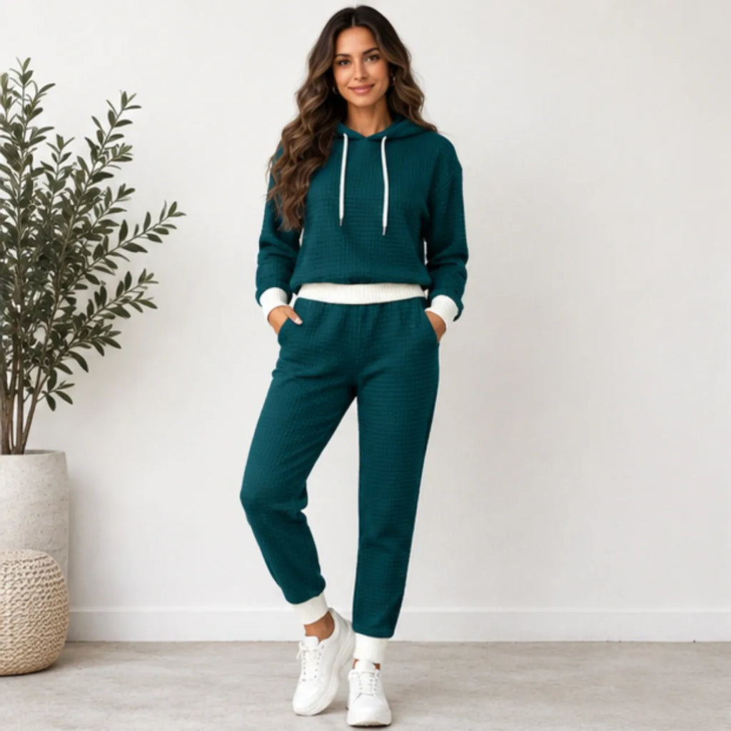 A woman modeling a matching two-piece lounge set consisting of a dark teal waffle-knit hoodie and jogger pants with white ribbed trim and white sneakers, standing in a brightly lit room next to a potted plant.
