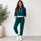 A woman modeling a matching two-piece lounge set consisting of a dark teal waffle-knit hoodie and jogger pants with white ribbed trim and white sneakers, standing in a brightly lit room next to a potted plant.