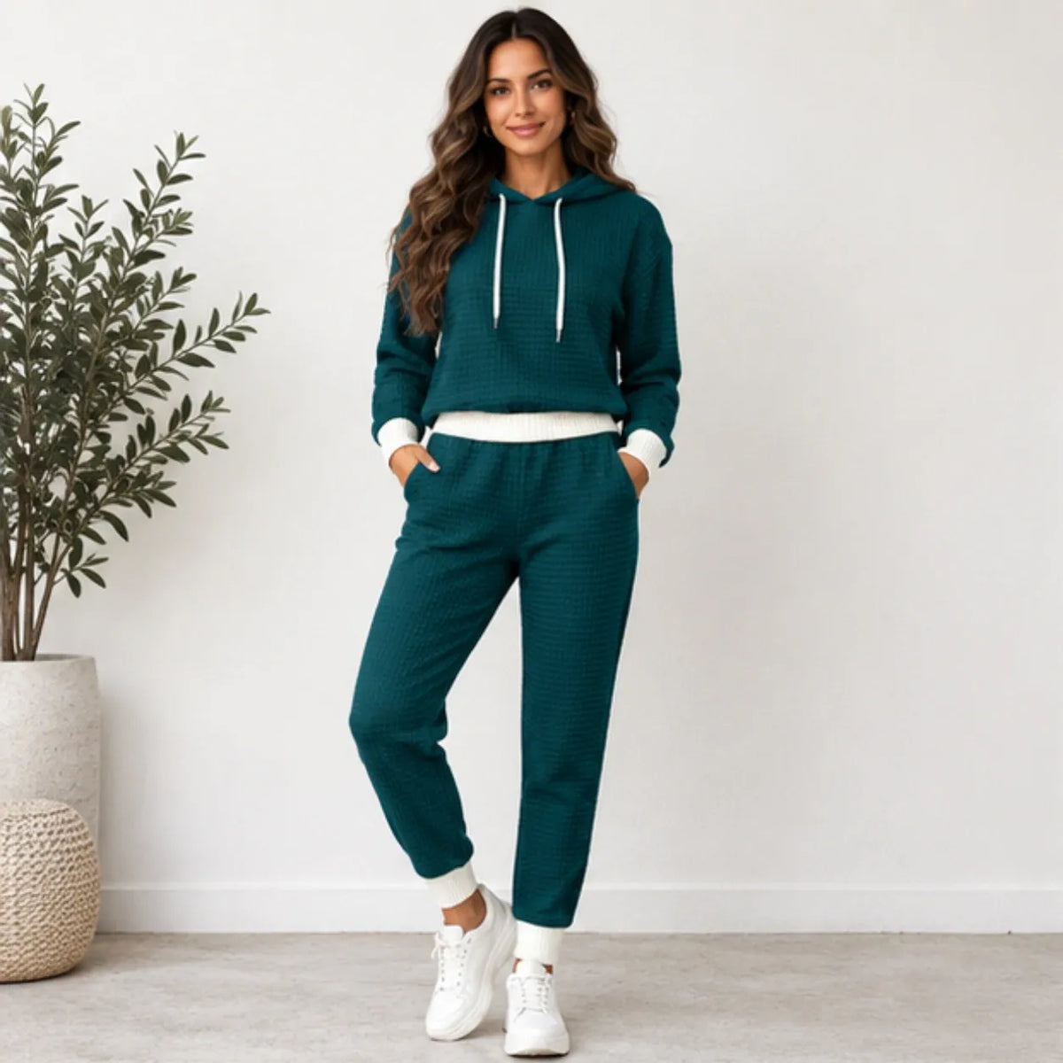 A woman modeling a matching two-piece lounge set consisting of a dark teal waffle-knit hoodie and jogger pants with white ribbed trim and white sneakers, standing in a brightly lit room next to a potted plant.