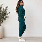 A woman modeling a matching two-piece waffle-knit loungewear set in deep teal, featuring a hooded sweatshirt and jogger pants, styled with white sneakers in a bright, modern room.