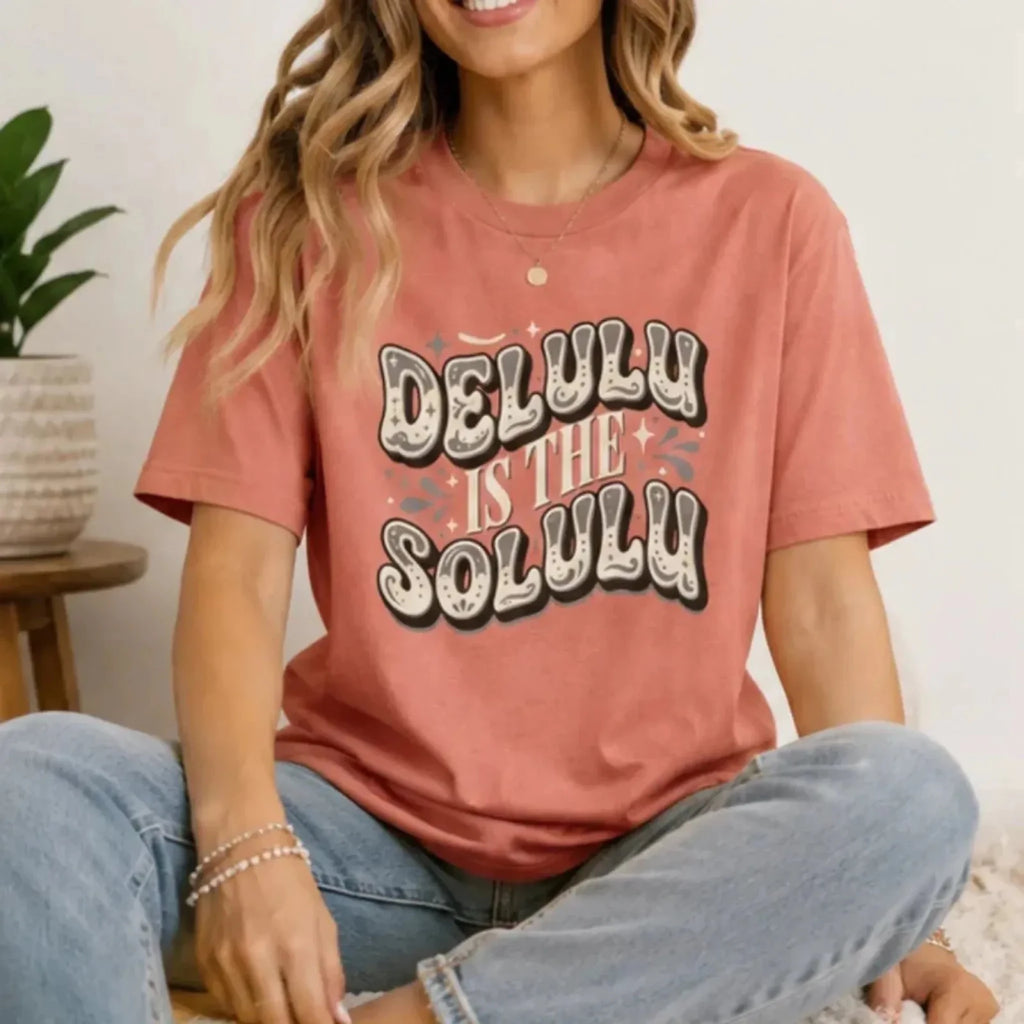 Woman sitting indoors wearing a dusty rose Delulu Is the Solulu graphic t-shirt with vintage-inspired statement graphic and casual denim jeans.