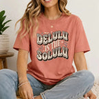 Woman sitting indoors wearing a dusty rose Delulu Is the Solulu graphic t-shirt with vintage-inspired statement graphic and casual denim jeans.