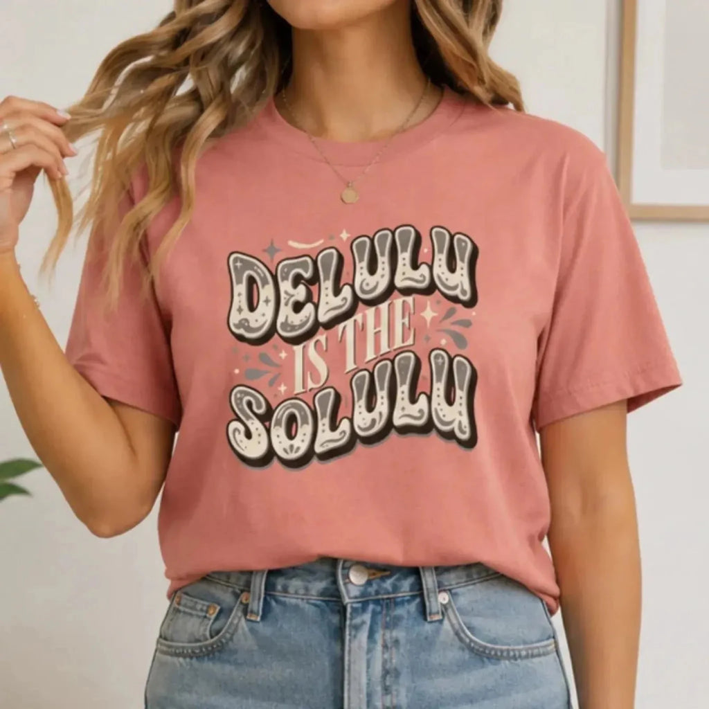 Woman wearing a dusty rose Delulu Is the Solulu graphic tee with retro inspirational typography paired with light wash denim jeans.