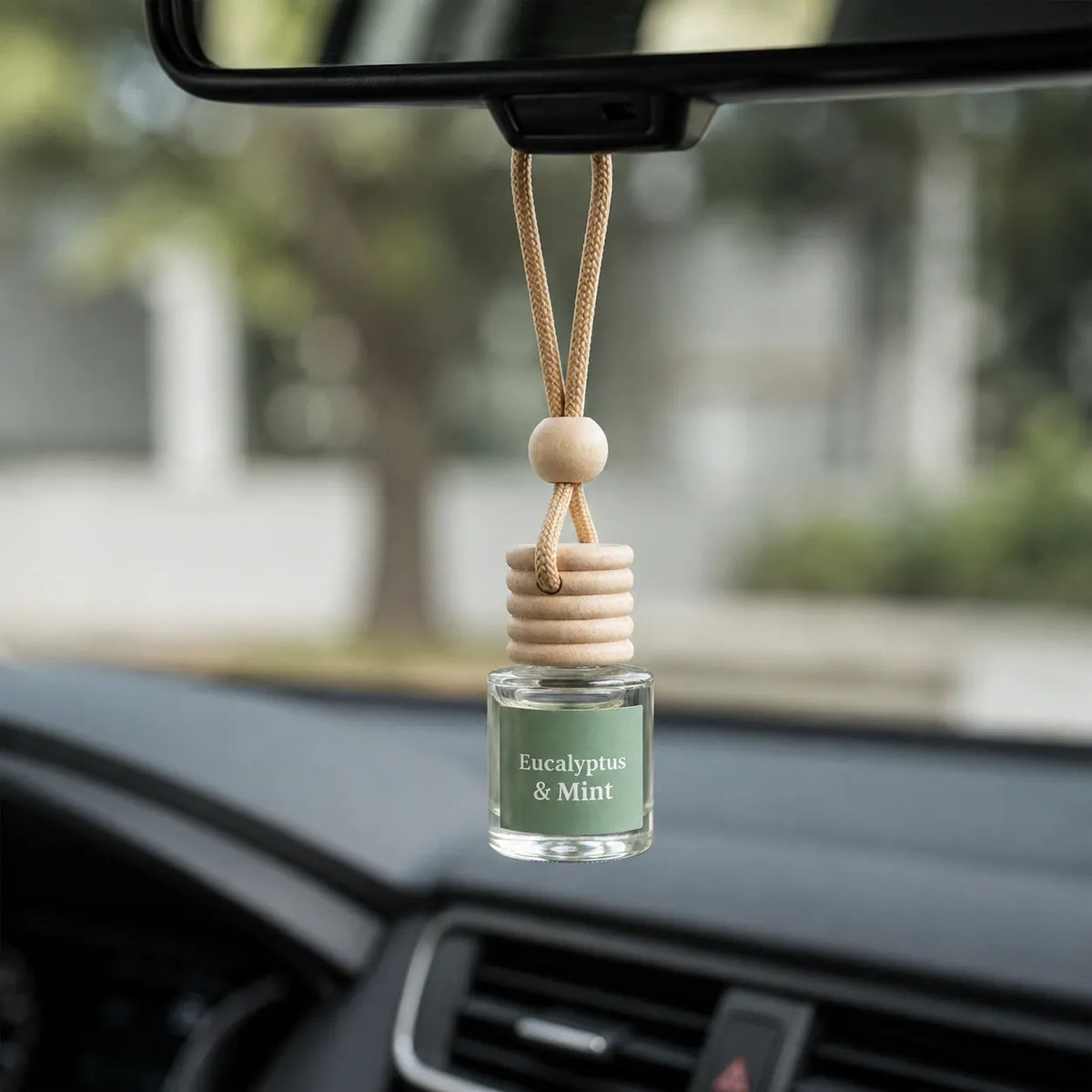 A car air freshener bottle labeled 'Eucalyptus & Mint' hanging from a car's rearview mirror. The bottle has a light green liquid and a natural wood diffuser top with a wooden bead.