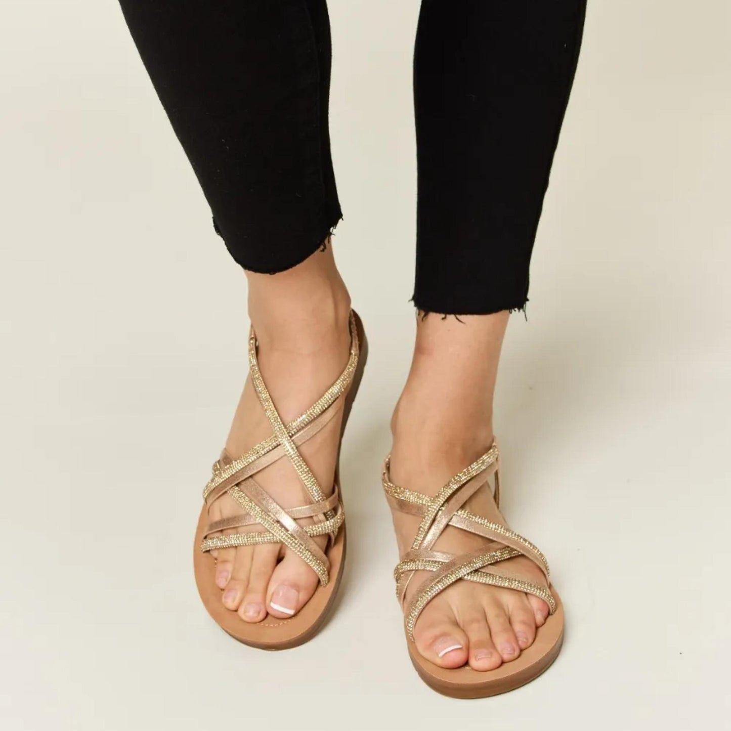 Front view of Forever Link gold rhinestone crisscross flat sandals worn with black trousers.