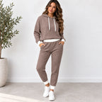 A woman modeling a matching two-piece lounge set consisting of a waffle-knit cropped hoodie and jogger pants in taupe brown, styled with white drawstrings, a white undershirt, and white sneakers.