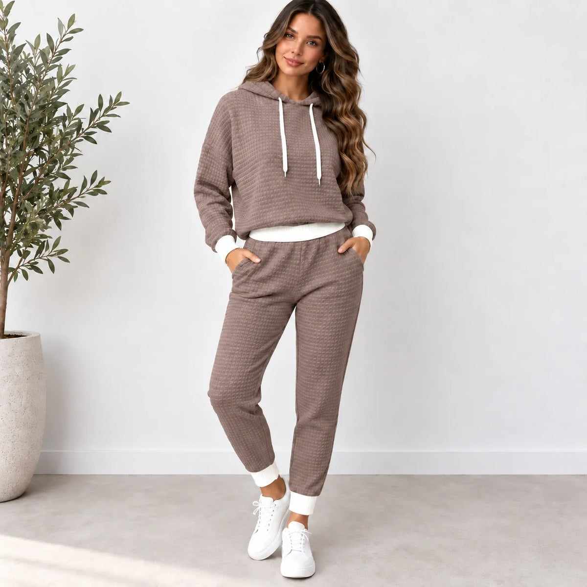 A woman modeling a matching two-piece lounge set consisting of a waffle-knit cropped hoodie and jogger pants in taupe brown, styled with white drawstrings, a white undershirt, and white sneakers.