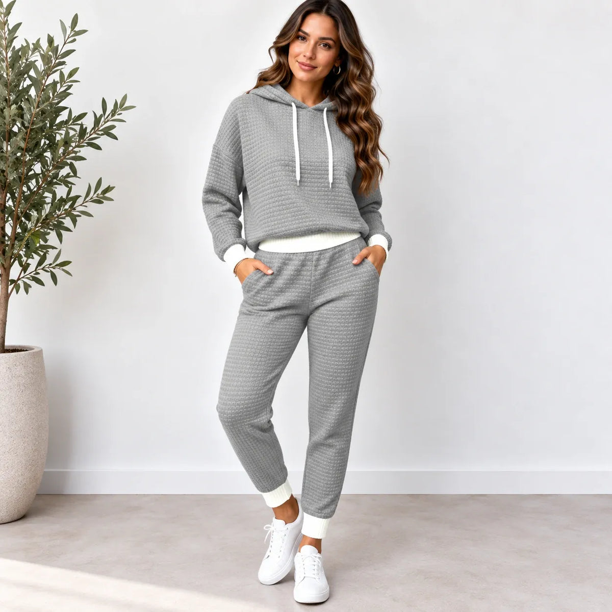 A woman modeling a matching gray waffle-knit lounge set consisting of a pullover hoodie and jogger pants, styled with white drawstrings, white ribbed trim, and white sneakers.