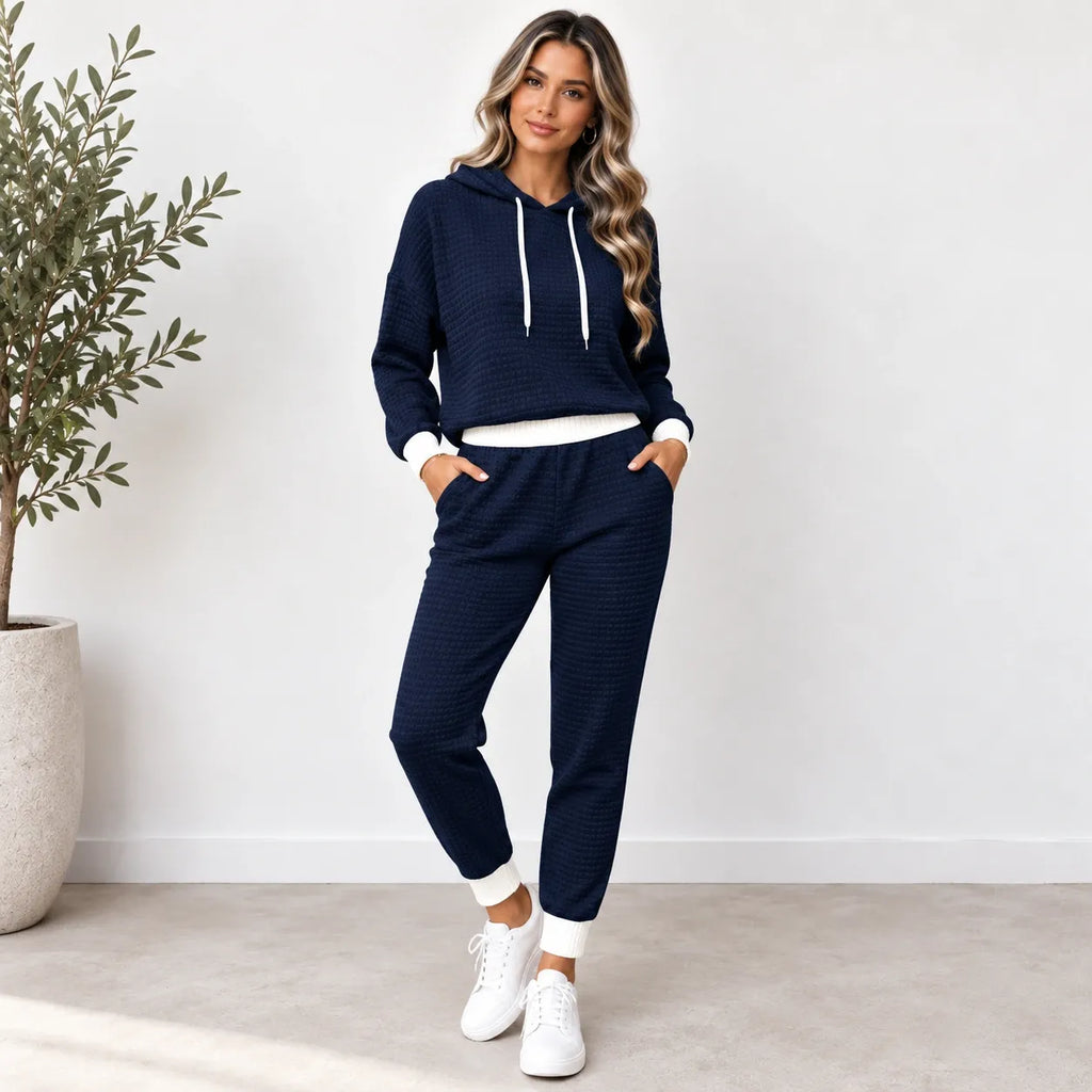 A woman modeling a matching navy blue textured waffle-knit lounge set, consisting of a pullover hoodie with white drawstrings and white ribbed trim, paired with matching jogger pants with white cuffs, styled with white sneakers in a bright indoor setting.