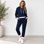A woman modeling a matching navy blue textured waffle-knit lounge set, consisting of a pullover hoodie with white drawstrings and white ribbed trim, paired with matching jogger pants with white cuffs, styled with white sneakers in a bright indoor setting.