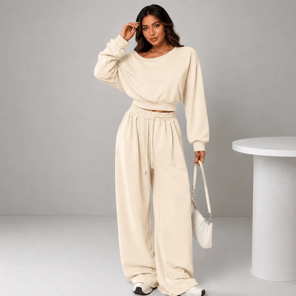 A woman modeling a matching cream-colored lounge set consisting of a cropped long-sleeve sweatshirt and wide-leg sweatpants, styled with a white handbag and sneakers against a neutral gray studio background.