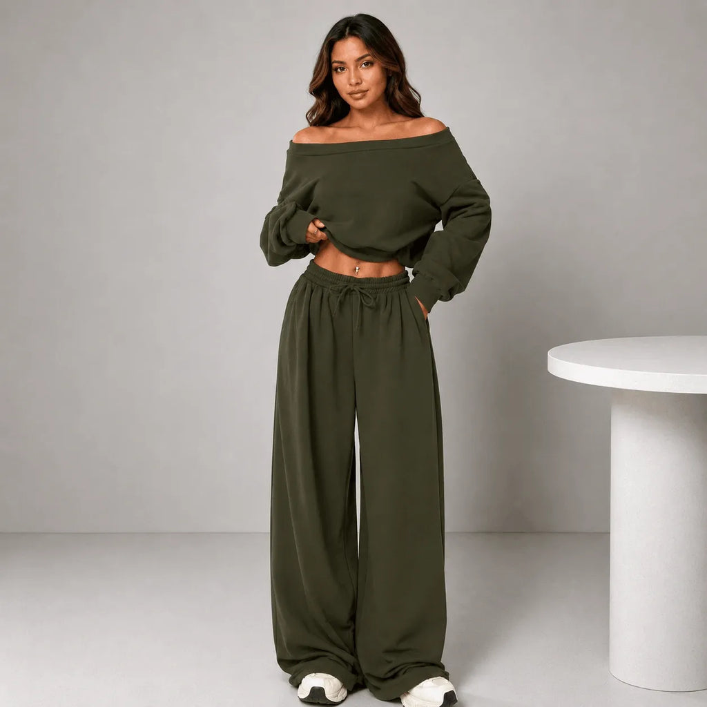 A woman modeling an olive green two-piece loungewear set consisting of an off-the-shoulder long-sleeve crop top and matching wide-leg drawstring pants, standing in a studio setting next to a white table.