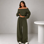 A woman modeling an olive green two-piece loungewear set consisting of an off-the-shoulder long-sleeve crop top and matching wide-leg drawstring pants, standing in a studio setting next to a white table.