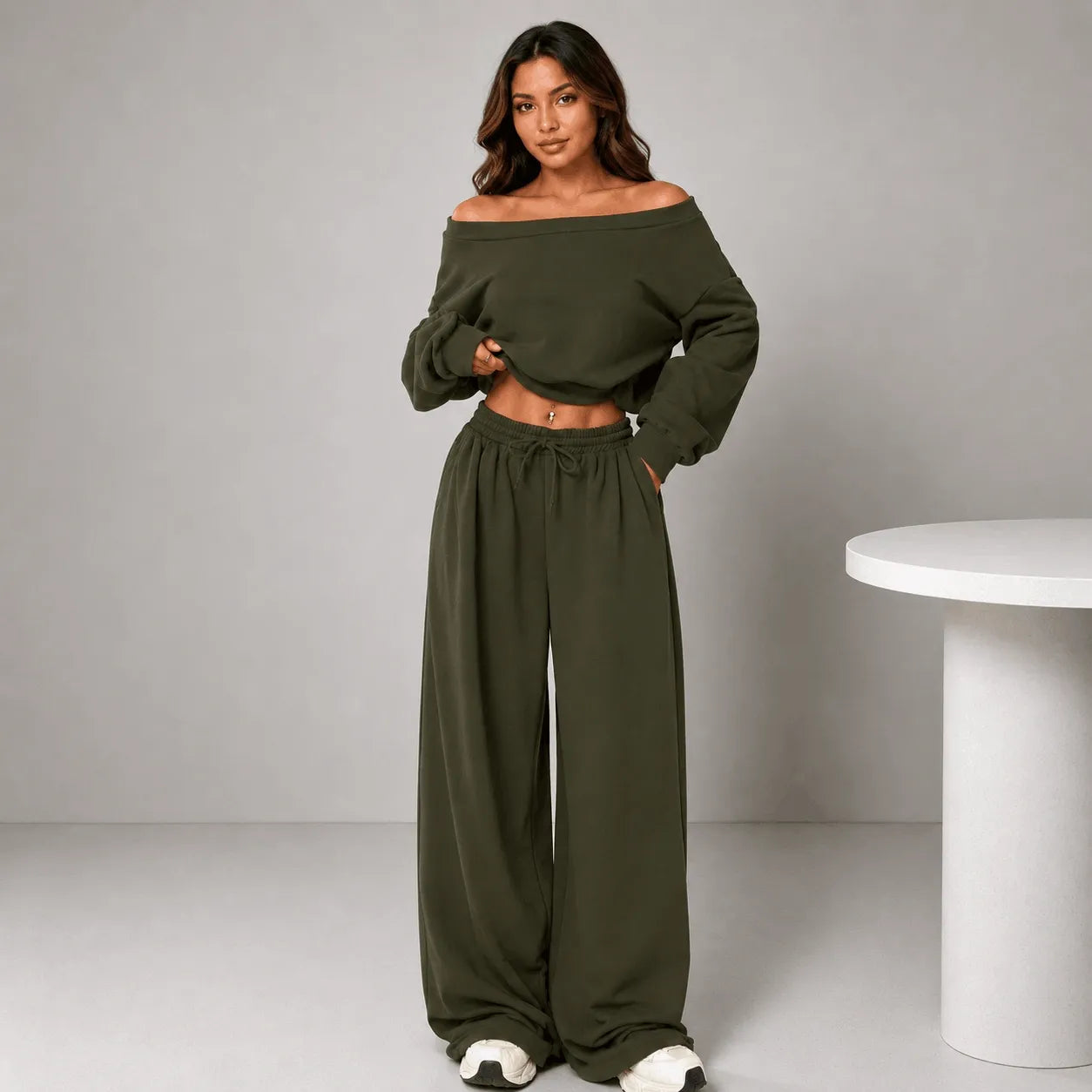 A woman modeling an olive green two-piece loungewear set consisting of an off-the-shoulder long-sleeve crop top and matching wide-leg drawstring pants, standing in a studio setting next to a white table.