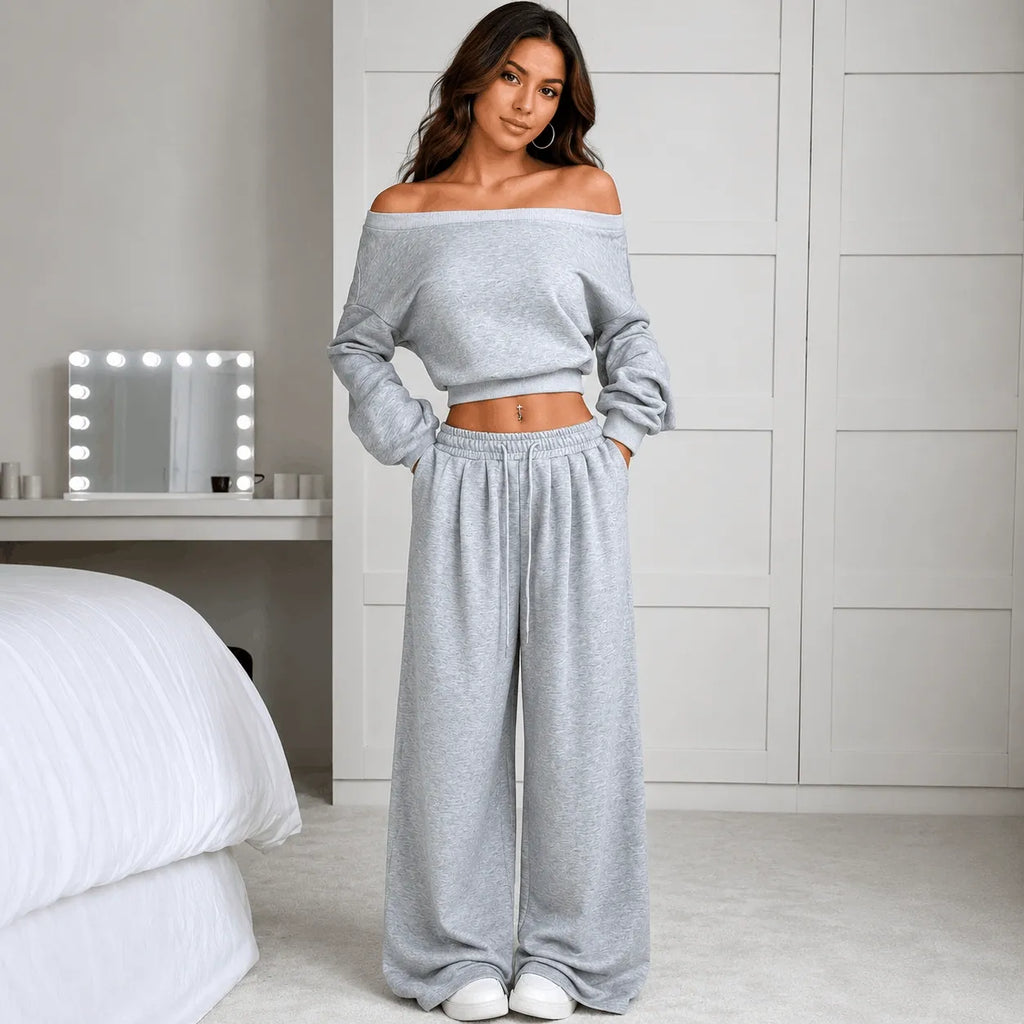 A woman stands in a bedroom wearing a light gray off-the-shoulder cropped sweatshirt and matching wide-leg sweatpants. The room has white walls, a bed with white bedding, and a vanity mirror with light bulbs.