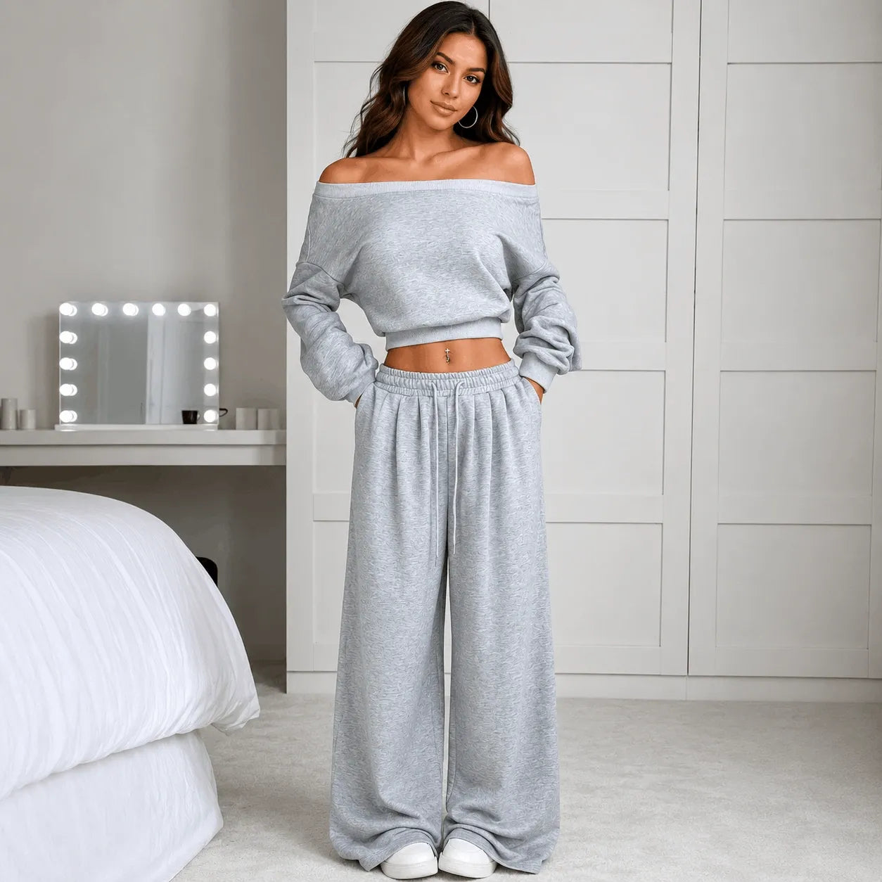A woman stands in a bedroom wearing a light gray off-the-shoulder cropped sweatshirt and matching wide-leg sweatpants. The room has white walls, a bed with white bedding, and a vanity mirror with light bulbs.