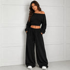 A woman modeling a black off-the-shoulder cropped long-sleeve top paired with matching black wide-leg sweatpants, standing against a light gray paneled wall.