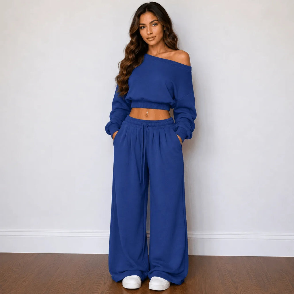 A woman modeling a matching royal blue loungewear set consisting of an off-the-shoulder long-sleeve crop top and wide-leg drawstring pants, paired with white sneakers, standing against a plain white wall.
