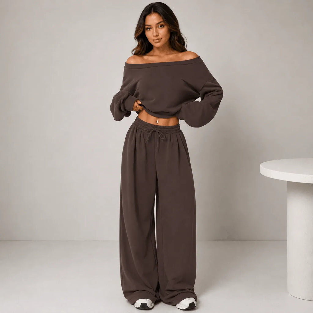 A woman modeling a matching dark brown loungewear set consisting of an off-the-shoulder long-sleeve crop top and high-waisted wide-leg drawstring pants, set against a minimalist studio background with a white table on the right.