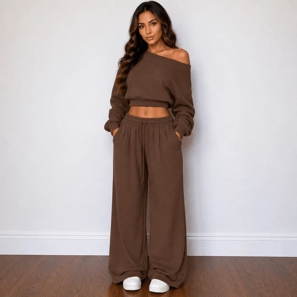 A woman modeling a matching brown two-piece loungewear set consisting of an off-the-shoulder long-sleeve crop top and high-waisted wide-leg pants, standing against a plain white wall on a wooden floor.