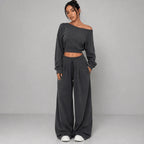 A woman modeling a dark gray two-piece loungewear set consisting of an off-the-shoulder long-sleeve crop top and matching wide-leg sweatpants, paired with white slip-on shoes against a light gray studio background.