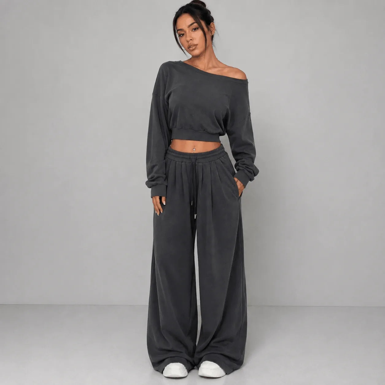 A woman modeling a dark gray two-piece loungewear set consisting of an off-the-shoulder long-sleeve crop top and matching wide-leg sweatpants, paired with white slip-on shoes against a light gray studio background.