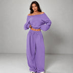 A woman modeling a matching lavender off-the-shoulder long-sleeve crop top and wide-leg drawstring sweatpants against a neutral gray studio background.