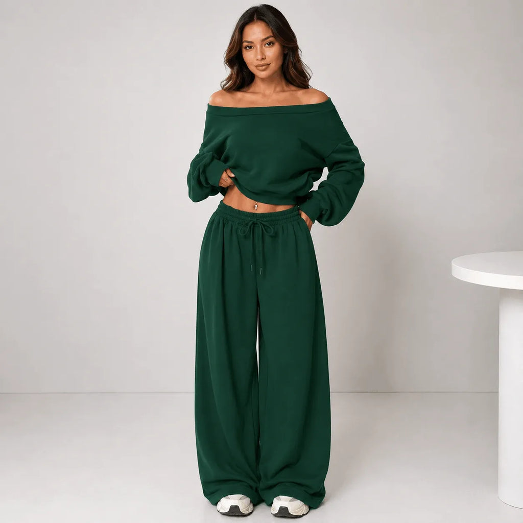 A woman modeling a dark green two-piece loungewear set consisting of an off-the-shoulder long-sleeve crop top and matching wide-leg drawstring pants, standing against a light gray studio background.