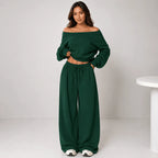 A woman modeling a dark green two-piece loungewear set consisting of an off-the-shoulder long-sleeve crop top and matching wide-leg drawstring pants, standing against a light gray studio background.