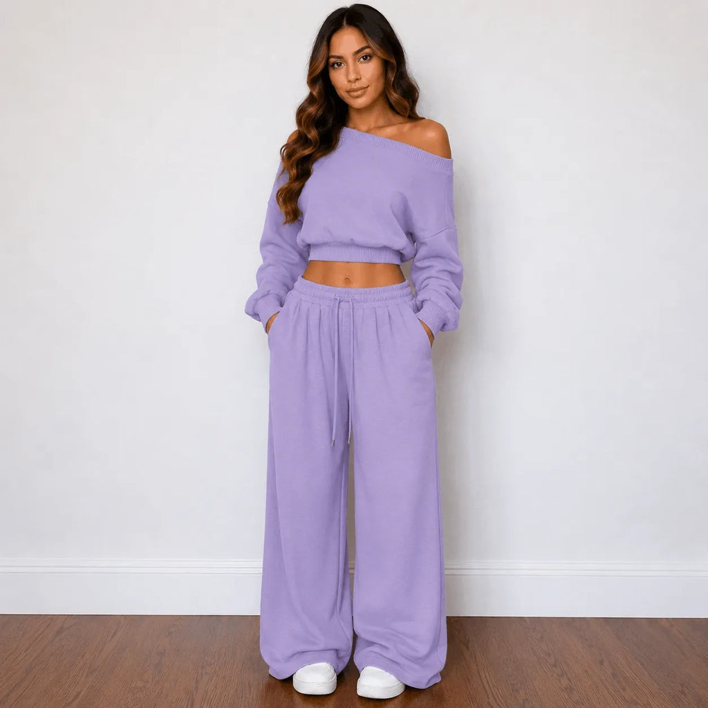 A woman modeling a matching lavender two-piece lounge set consisting of an off-the-shoulder long-sleeve crop top and wide-leg drawstring pants, standing against a white wall on a wooden floor.