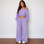 A woman modeling a matching lavender two-piece lounge set consisting of an off-the-shoulder long-sleeve crop top and wide-leg drawstring pants, standing against a white wall on a wooden floor.