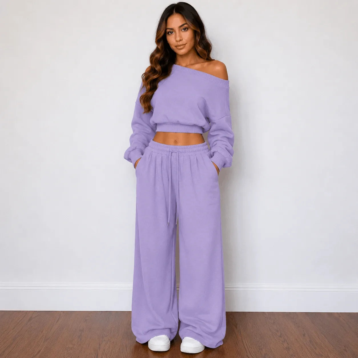 A woman modeling a matching lavender two-piece lounge set consisting of an off-the-shoulder long-sleeve crop top and wide-leg drawstring pants, standing against a white wall on a wooden floor.