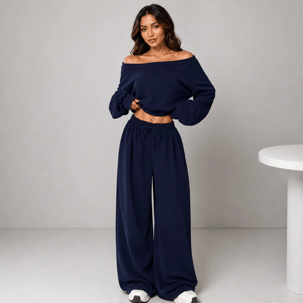 A woman modeling a navy blue two-piece loungewear set consisting of an off-the-shoulder long-sleeve crop top and matching wide-leg drawstring pants, standing against a light gray studio background.