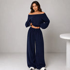 A woman modeling a navy blue two-piece loungewear set consisting of an off-the-shoulder long-sleeve crop top and matching wide-leg drawstring pants, standing against a light gray studio background.