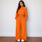 A woman modeling a bright orange two-piece lounge set consisting of an off-the-shoulder long-sleeve crop top and matching wide-leg drawstring pants, paired with white sneakers against a plain white wall and wood floor.
