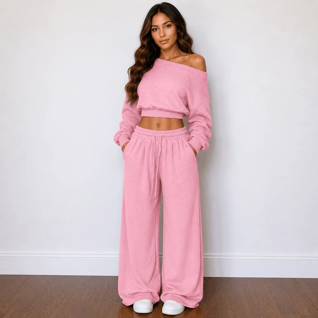 A woman modeling a matching pink two-piece lounge set consisting of an off-the-shoulder long-sleeve crop top and wide-leg drawstring pants, standing against a plain white wall on a wooden floor.