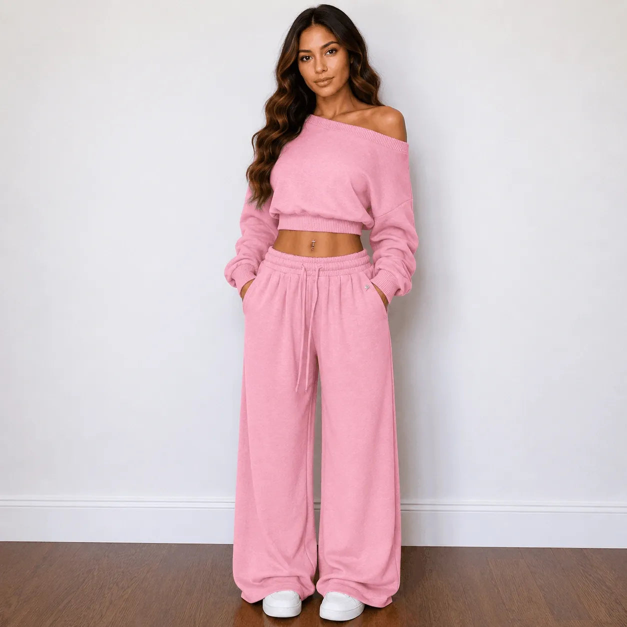 A woman modeling a matching pink two-piece lounge set consisting of an off-the-shoulder long-sleeve crop top and wide-leg drawstring pants, standing against a plain white wall on a wooden floor.
