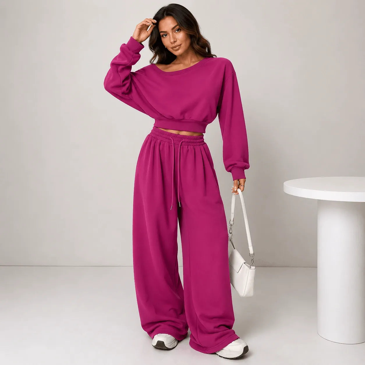 A woman models a matching two-piece lounge set in a bright magenta pink color, consisting of a cropped long-sleeve sweatshirt and high-waisted wide-leg sweatpants. She is standing in a studio setting next to a white cylindrical table.