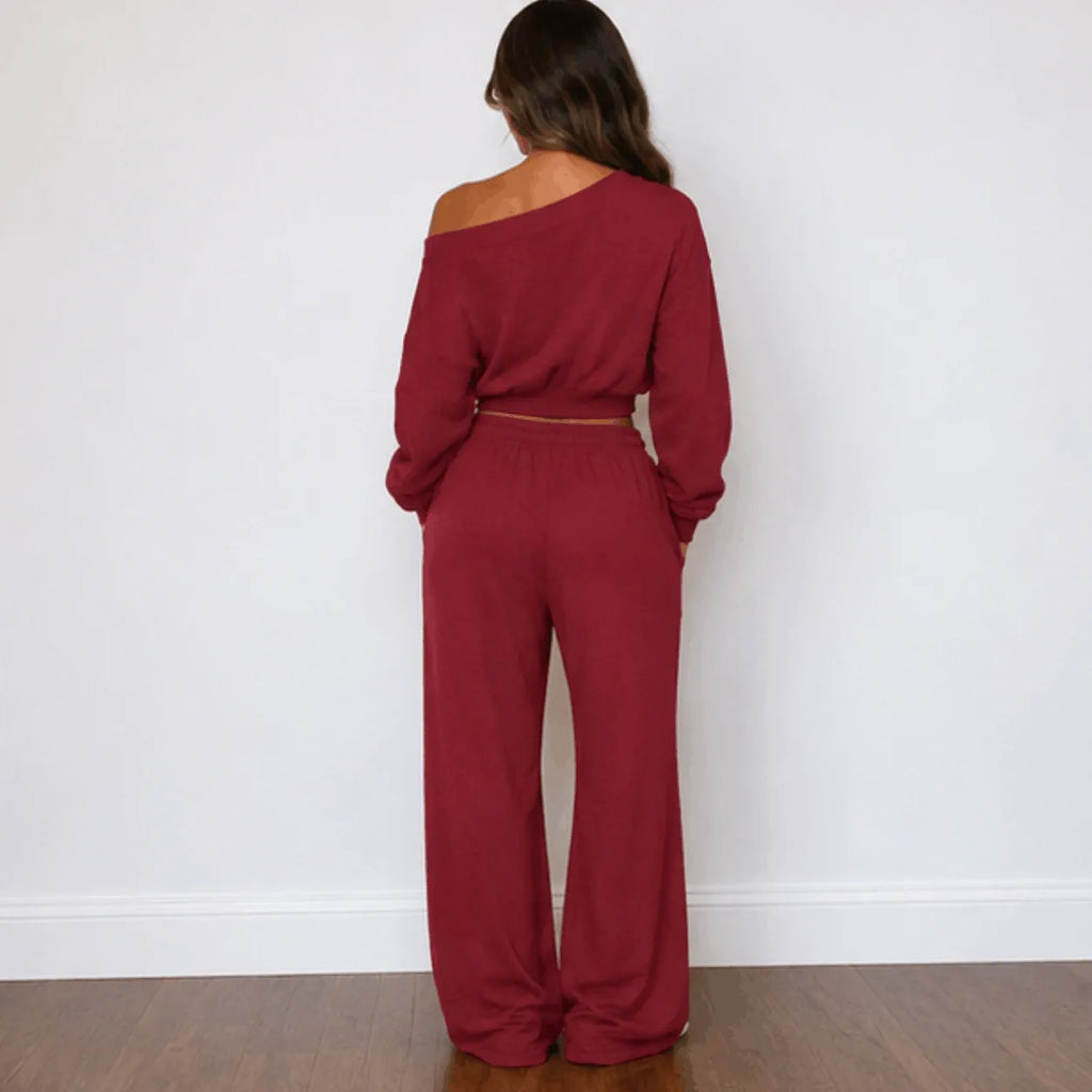 A woman stands with her back to the camera wearing a matching red two-piece loungewear set consisting of an off-the-shoulder long-sleeve crop top and wide-leg pants, set against a plain white wall and wooden floor.