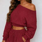 A woman wearing a long-sleeve off-the-shoulder cropped knit sweater in red, paired with matching jogger pants, against a plain light gray background.