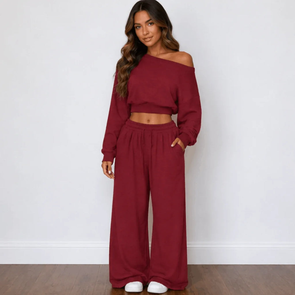 A woman modeling a matching red two-piece loungewear set consisting of an off-the-shoulder long-sleeve crop top and high-waisted wide-leg pants, paired with white slip-on shoes against a white wall and wood floor.
