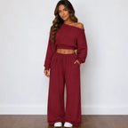 A woman modeling a matching red two-piece loungewear set consisting of an off-the-shoulder long-sleeve crop top and high-waisted wide-leg pants, paired with white slip-on shoes against a white wall and wood floor.